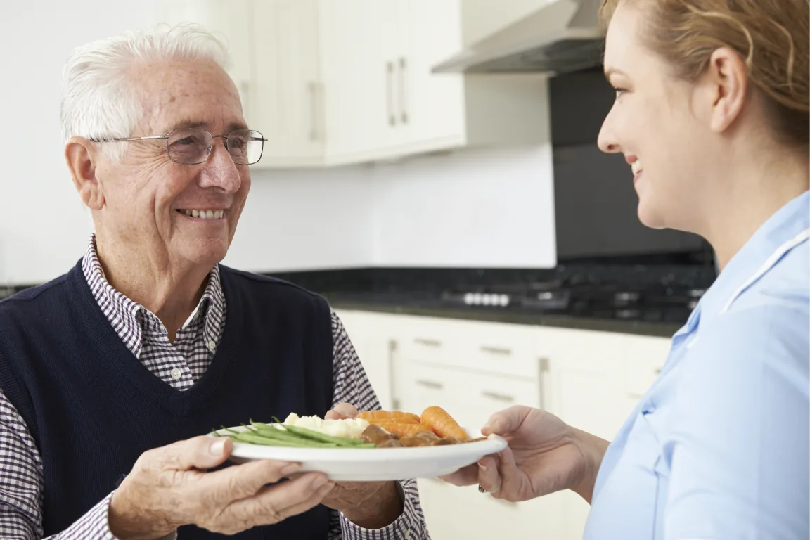 A young woman handing a warm meal to a smiling elderly man in a kitchen.